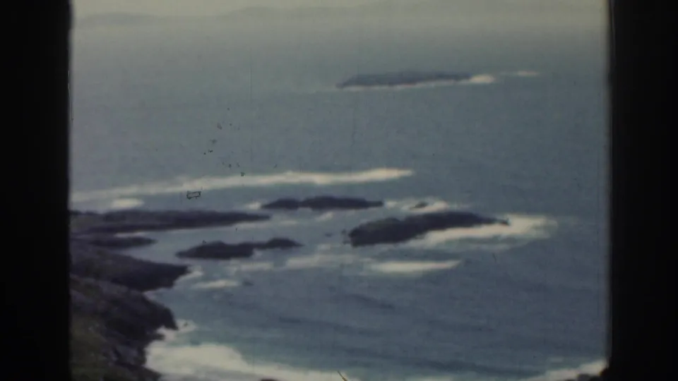1961 Ireland 8mm frame showing hazy ocean, rocky outcrops, and surf viewed through a dark window mullion.