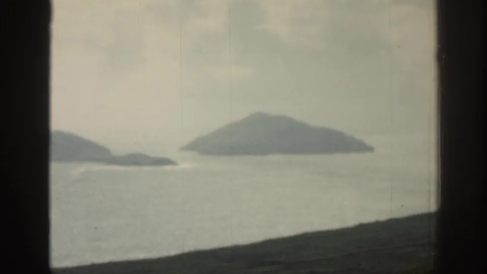 1961 Ireland coastal frame with a mist-softened island, pale sea, and dark foreground land viewed through glass.