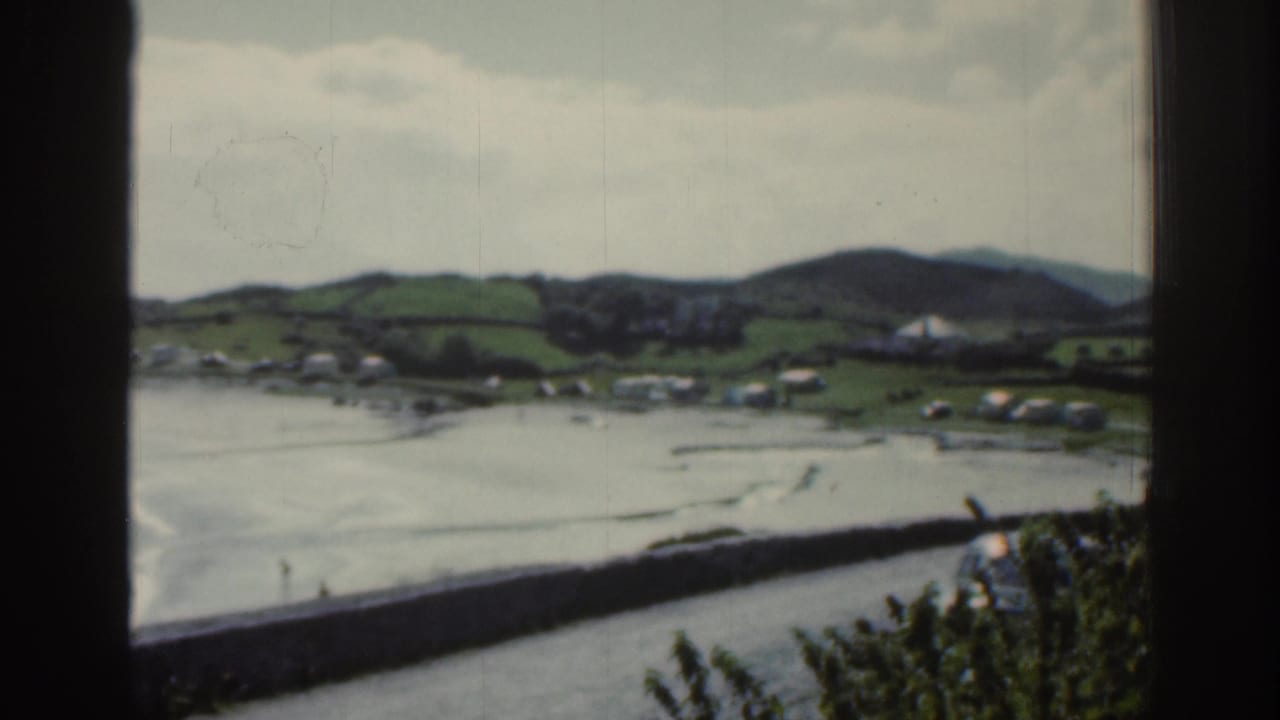1961 Ireland 8mm frame of a rural bay settlement with green hills, scattered white cottages, cloudy sky, and a long seawall.