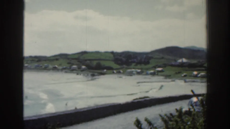1961 Ireland home-movie view of a shoreline village, green hills, bay water, and seawall divided by dark foreground framing.