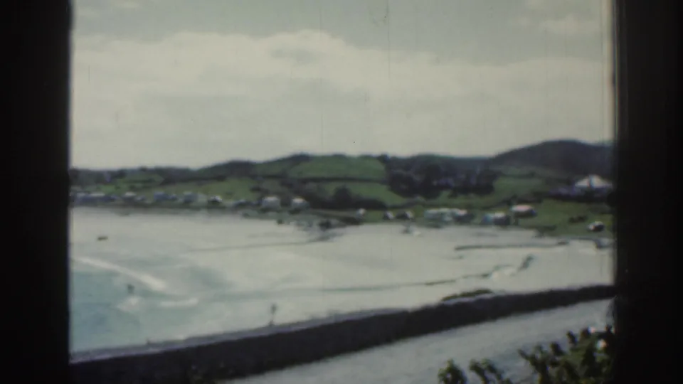 1961 Ireland coastal frame showing an overcast bay, small shoreline settlement, rolling hills, a seawall, and foliage in the foreground.