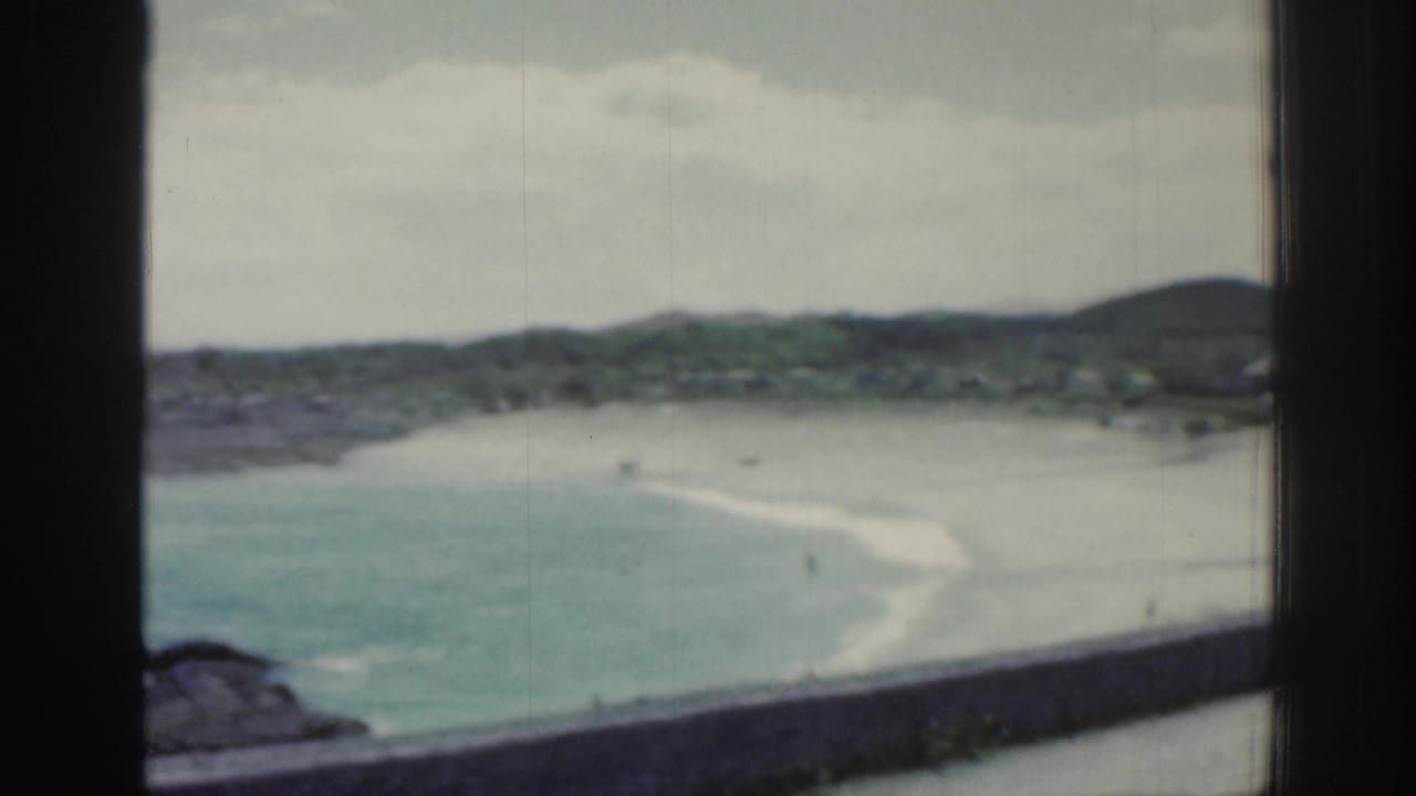 1961 Ireland home-movie view of a sandy bay through a window frame, with blue-green water, rocky shore, low hills, and distant beach activity.