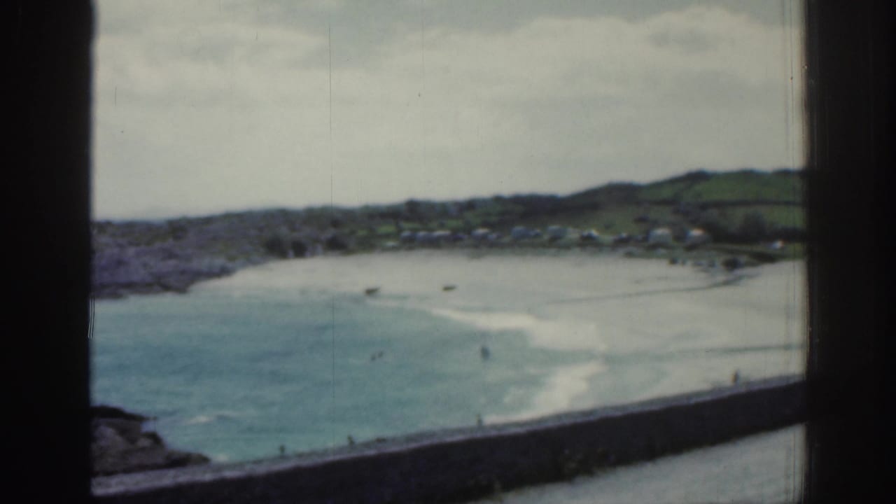 1961 Ireland coastal scene with a sandy beach, blue-green shallows, distant white caravans, low green hills, and small figures by the water.
