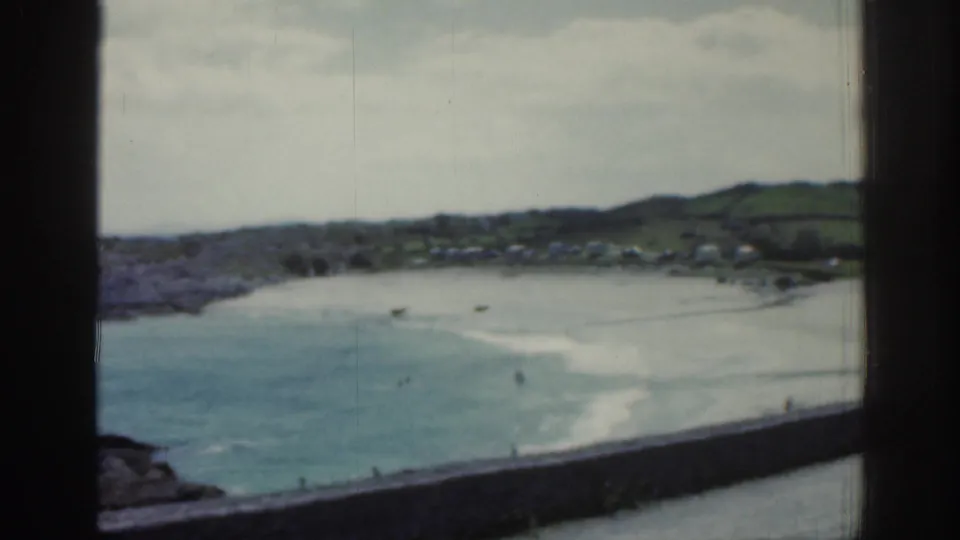 1961 Ireland coastal scene with a sandy beach, blue-green shallows, distant white caravans, low green hills, and small figures by the water.