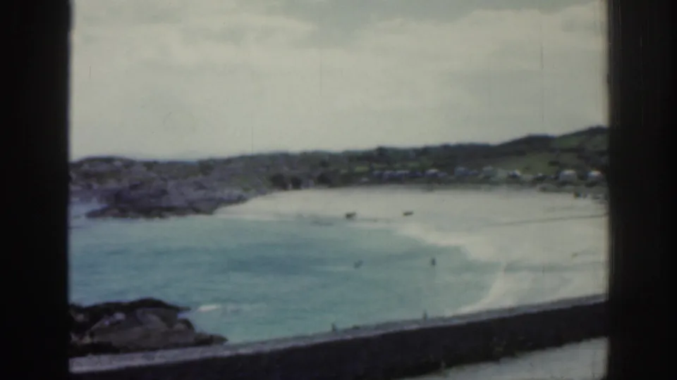 Soft 8mm view of an Irish beach bay in 1961, framed by dark window bars with shallow water, pale sand, rocks, and distant people.