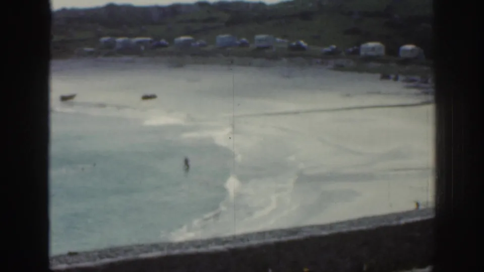 1961 Ireland beach detail with pale sandbars, shallow bay water, small boats, distant caravans, green hills, and tiny wading figures.