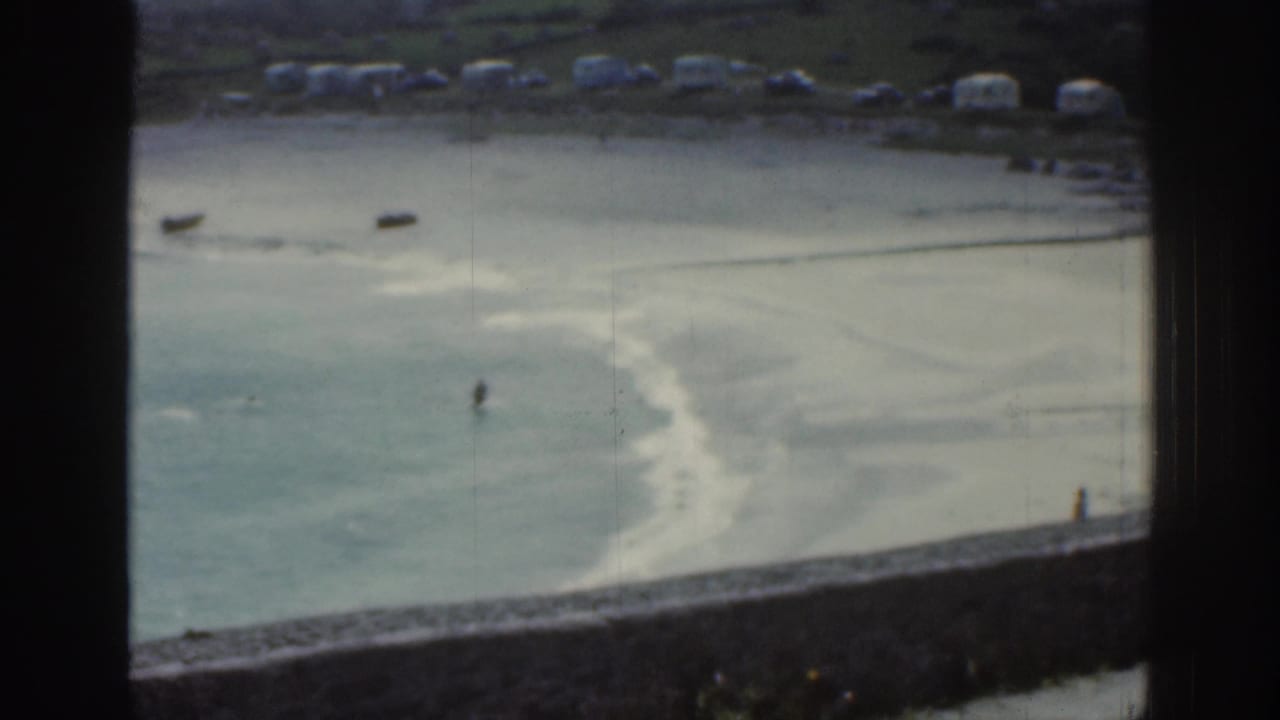 1961 Ireland coastal home-movie view through a window frame, showing shallow tidal water, exposed sand, and distant figures by the shore.