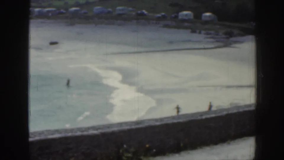1961 Ireland shoreline seen through a window, with pale sand, shallow channels, and small figures walking or wading near the water.