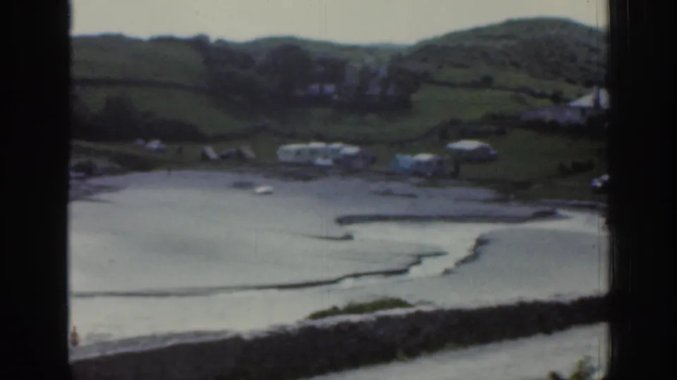 1961 Ireland home-movie frame showing a low-tide coastal inlet, grassy hillside, white caravans, small boats, and a dark window frame.