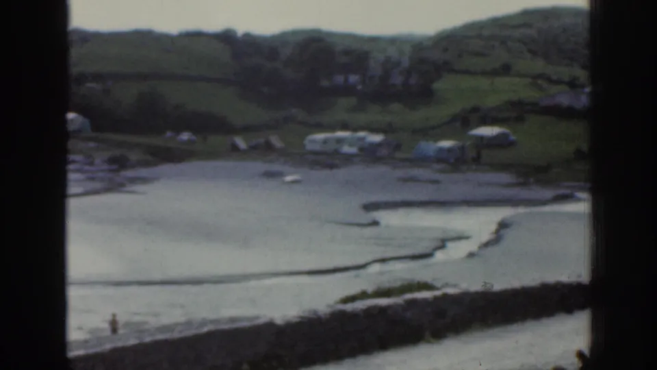 1961 Ireland coastal home-movie view of caravans above a low-tide beach, shallow channels, small boats, and a distant shoreline figure.