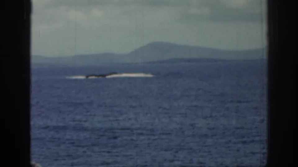 A 1961 Ireland coastal view shows distant white surf on rocks, open water, hazy hills, and heavy dark framing at both sides.