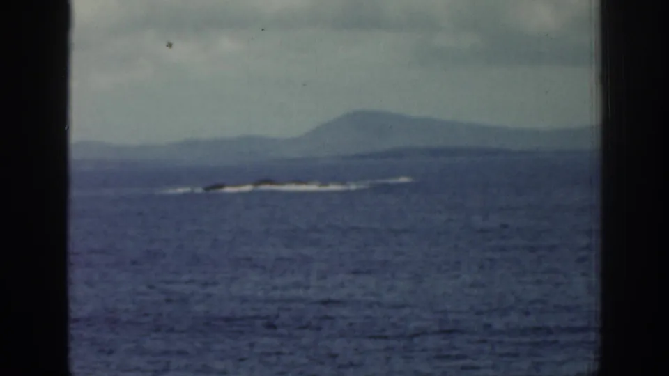The 8mm home-movie view looks across Irish coastal water toward low hills and a thin line of breaking waves in the distance.