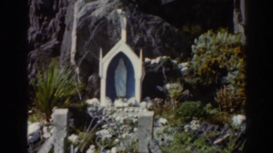 1961 Ireland home-movie frame showing a small white religious shrine with a blue figure below a rocky cliff and surrounding plants.