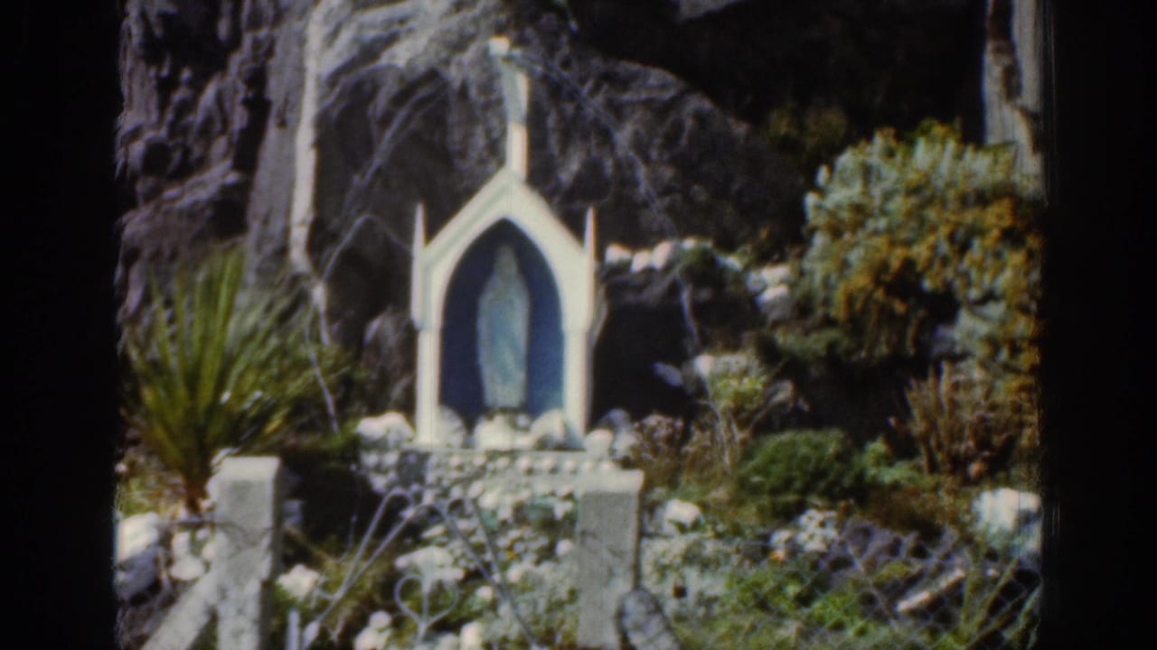 White shrine and blue figure framed by rocks, shrubs, and garden plants.