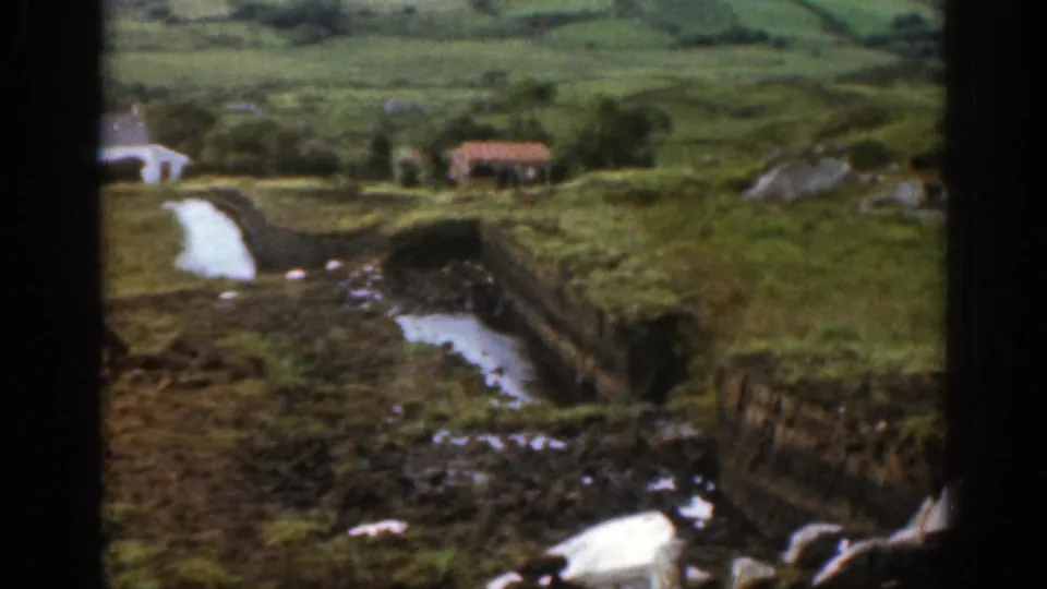 1961 Ireland countryside view showing a winding stream between green banks, with a small distant cottage and low hills beyond.
