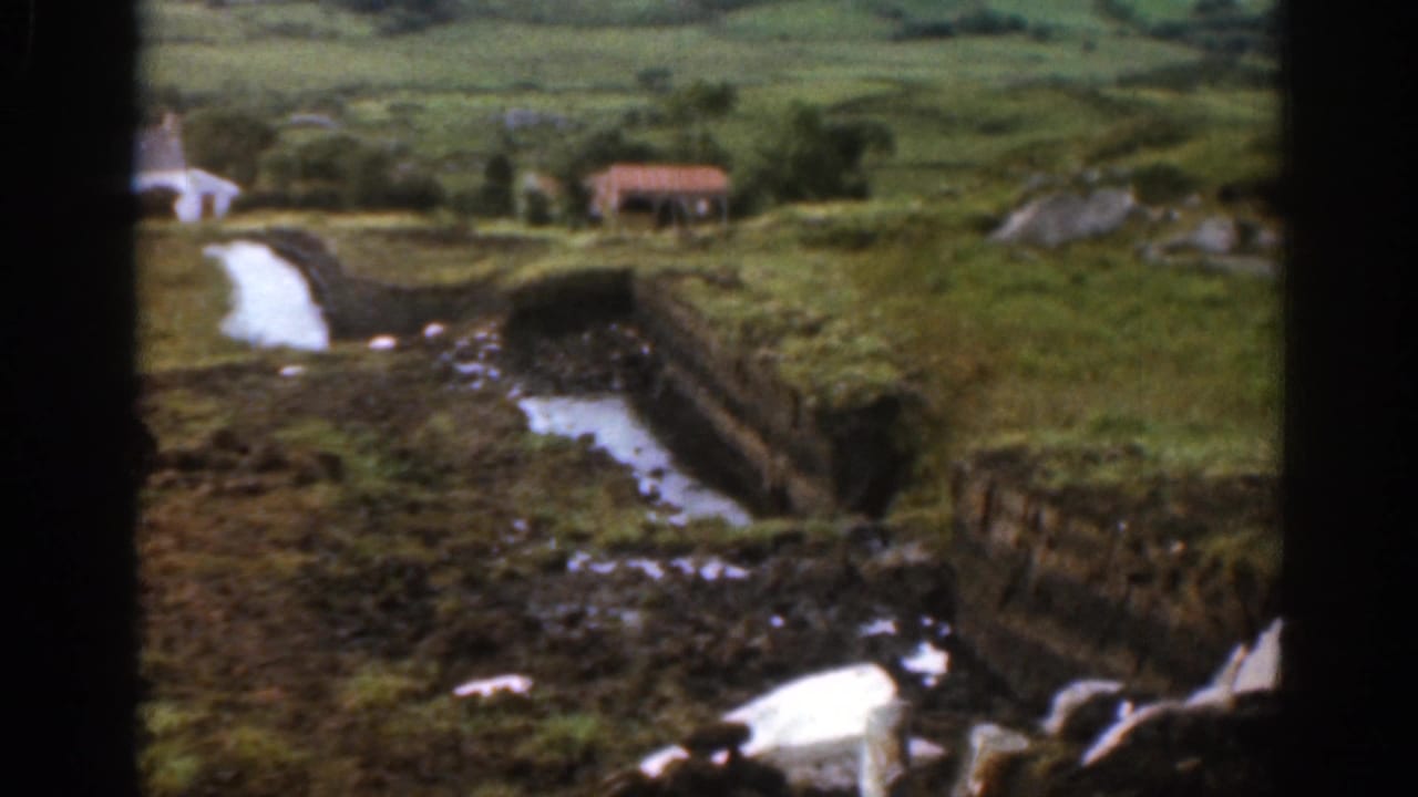 1961 Ireland home-movie frame showing the stream continuing through grassy fields toward small distant buildings and low hills.