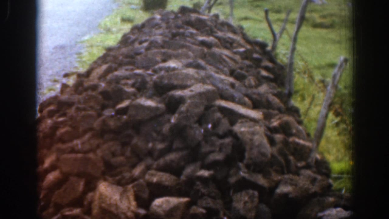 Close 1961 Ireland home-movie view along a weathered dry-stone wall with roadside grass and fence posts nearby.