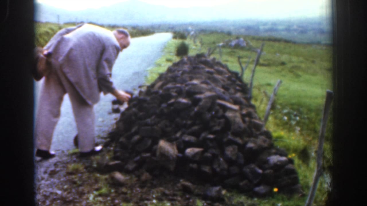 In 1961 Ireland, a man in a light-colored suit bends near a dry-stone wall beside a narrow road in a green rural valley.