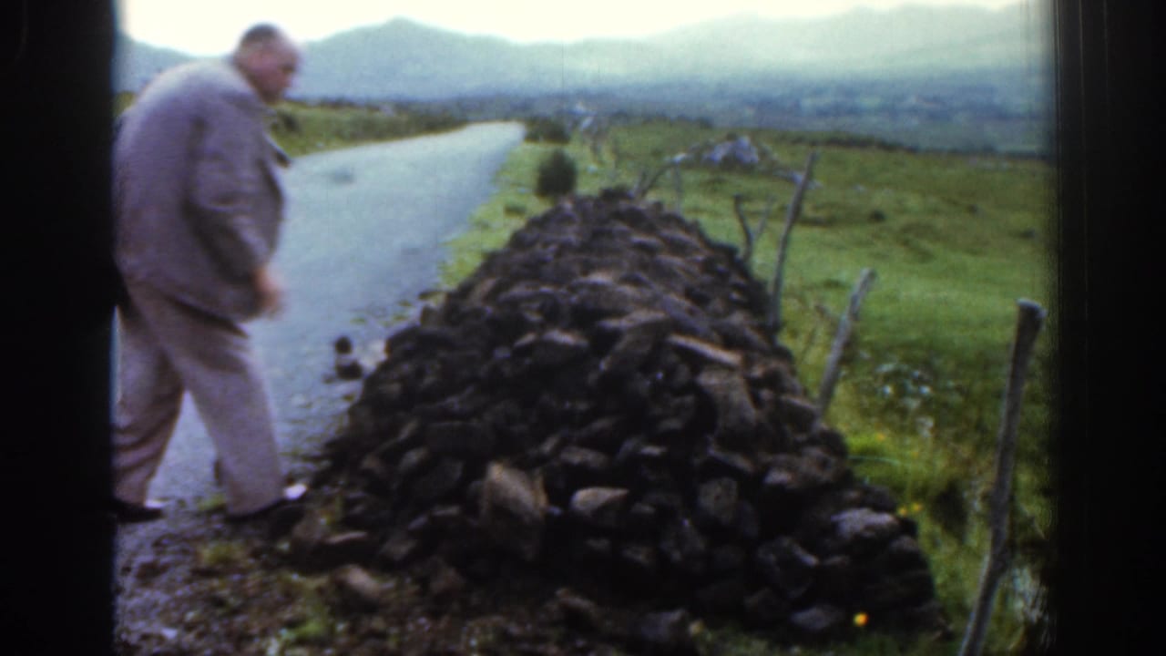 Man stands beside a stone wall along a country road with hills beyond.