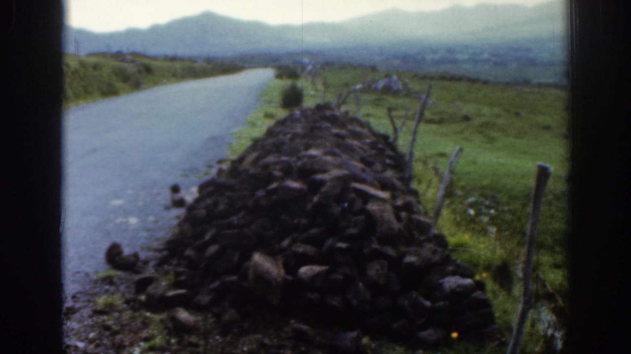 The camera holds on the stone wall beside the narrow road, with green Irish fields, fence posts, and hazy hills beyond.