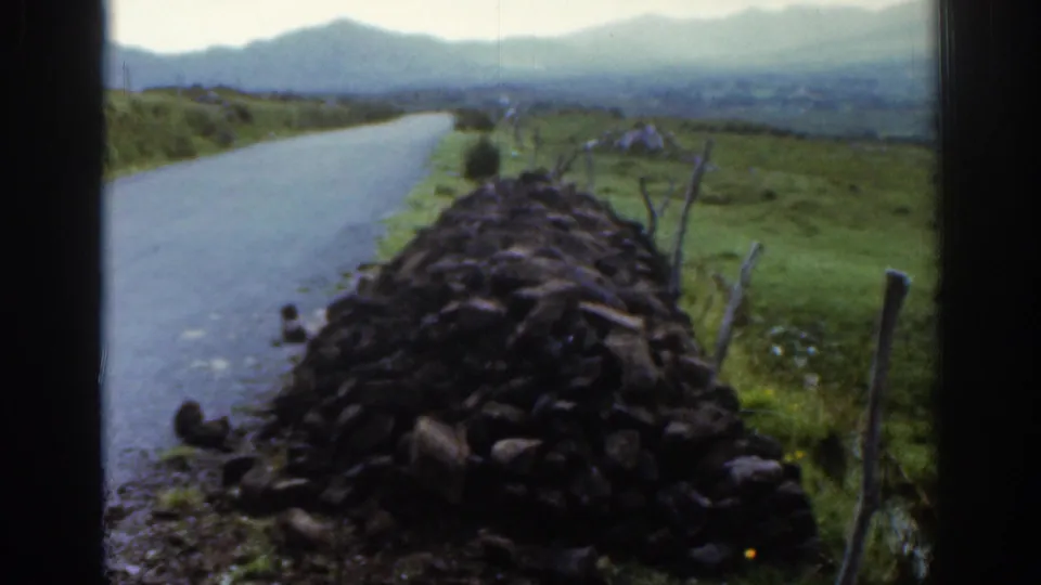 The camera holds on the stone wall beside the narrow road, with green Irish fields, fence posts, and hazy hills beyond.