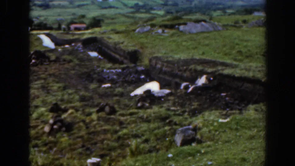 A wider rural Irish view shows stone boundary walls, green pasture, wet ground, scattered rocks, and small farm structures under cloudy light.