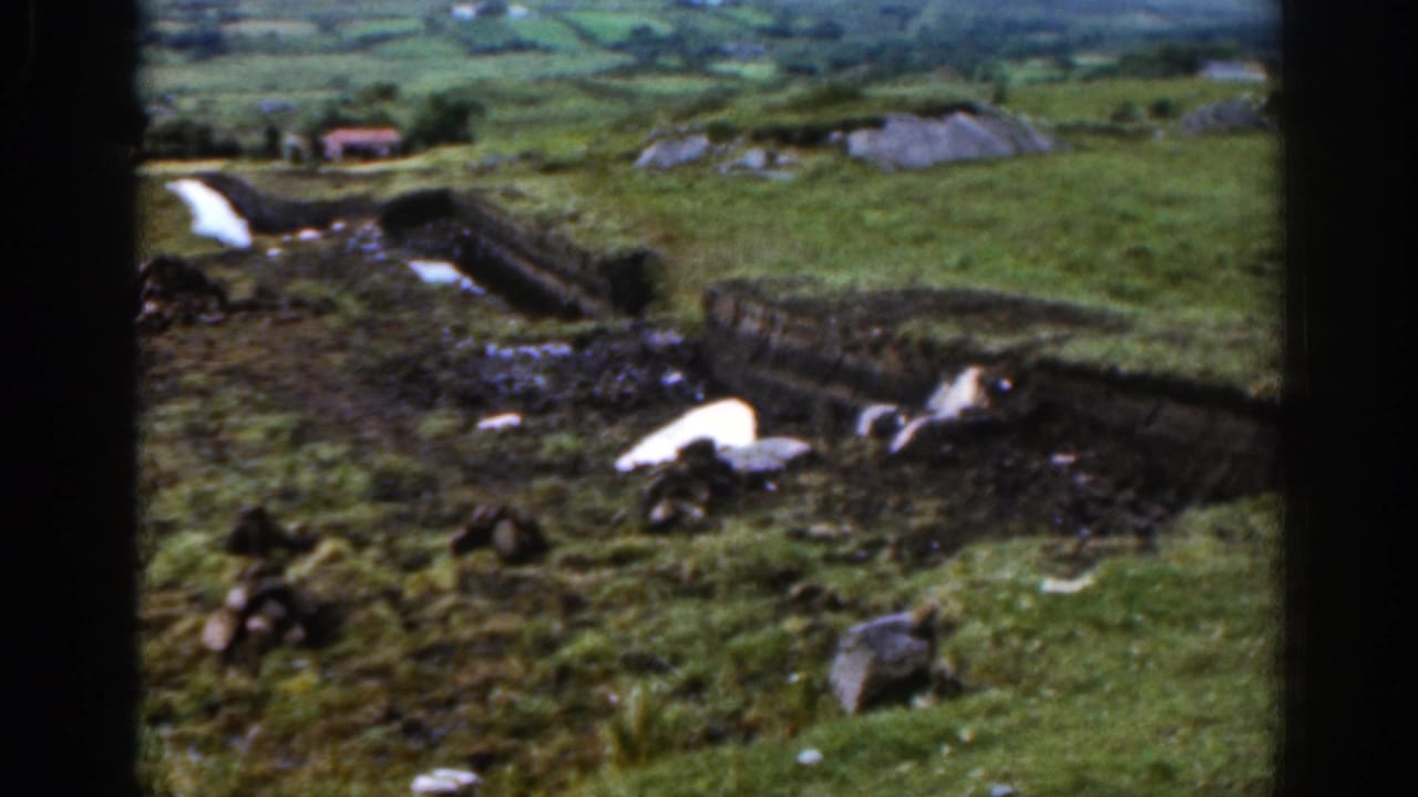 1961 Ireland home-movie frame showing a hillside farm area with low boundaries, scattered rocks, green fields, and distant valley farmland.