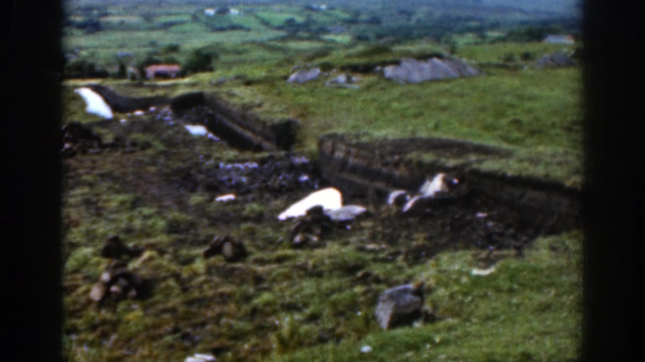 1961 Ireland countryside view with a hillside farm boundary, rocky ground, dark earth patches, and soft green fields stretching into the valley.