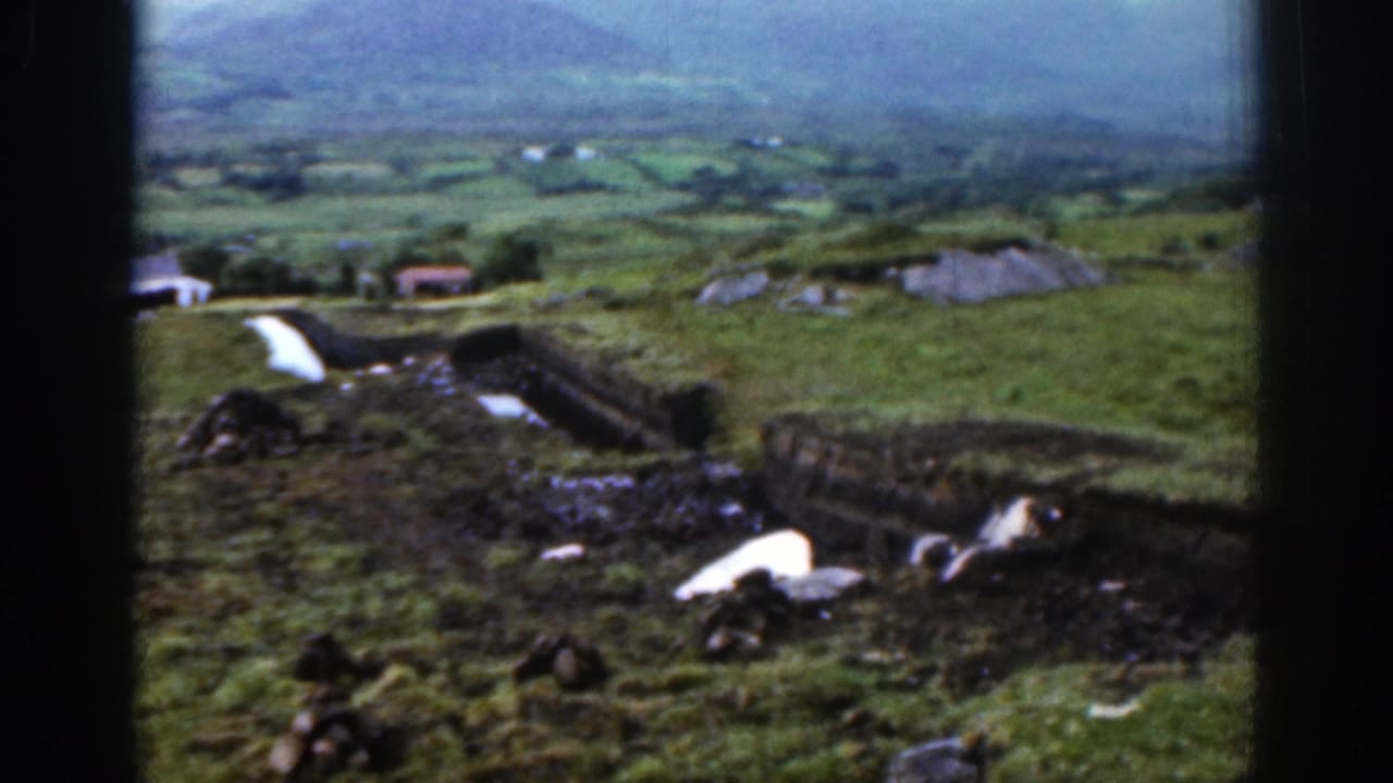 1961 Ireland 8mm frame showing a green valley, small farm buildings, a hillside boundary, and muted mountain light in the distance.