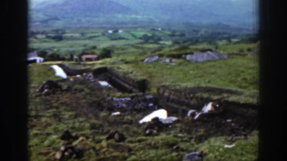 1961 Ireland 8mm frame showing a green valley, small farm buildings, a hillside boundary, and muted mountain light in the distance.