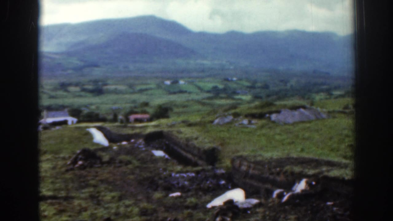 1961 Ireland home-movie view widening to show mountain slopes, valley fields, small farm buildings, and a rough hillside path or boundary.