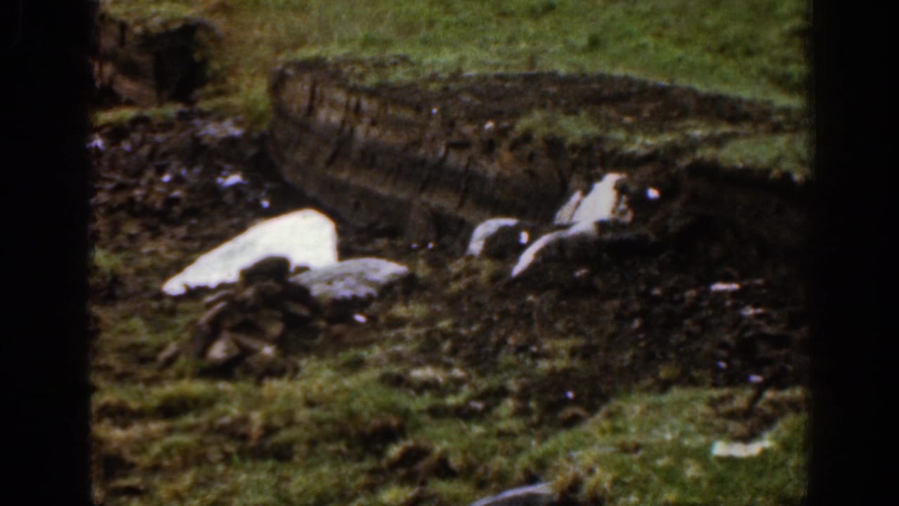 1961 Ireland 8mm frame showing melting snow or ice in a muddy grassy patch with rocks and a rough bank in the background.