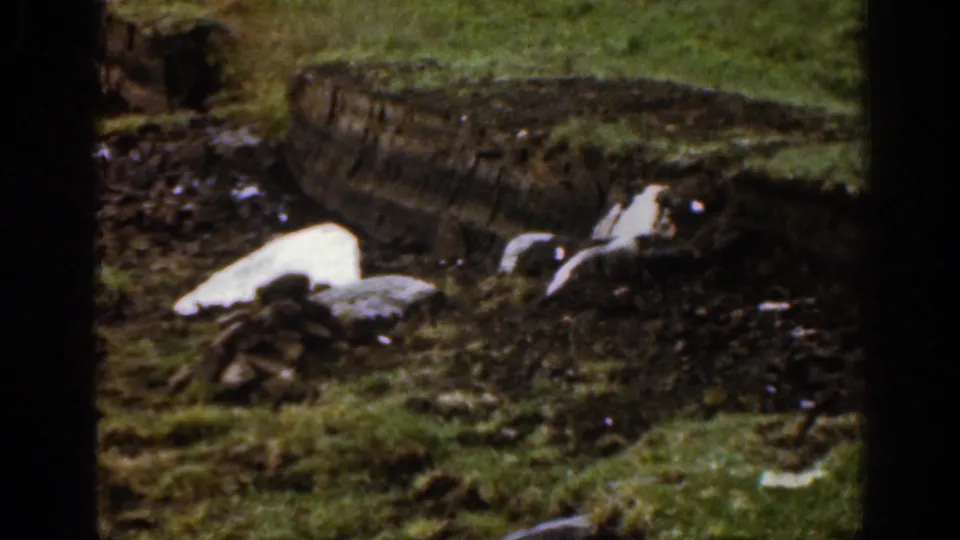 1961 Ireland 8mm frame showing melting snow or ice in a muddy grassy patch with rocks and a rough bank in the background.