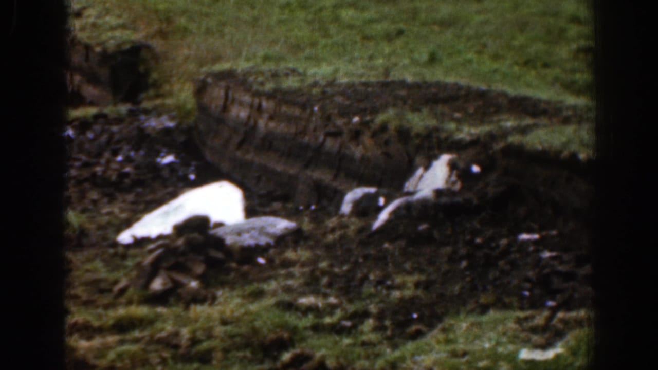 1961 Ireland 8mm view of a muddy rural patch with melting snow or ice, stones, grass, and a weathered bank at the rear.