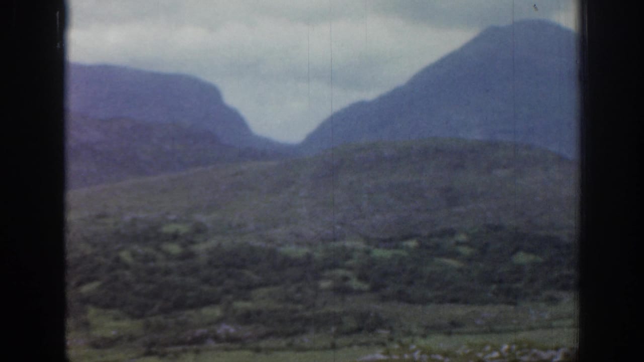 1961 Ireland 8mm frame showing green scrubby hills, distant blue-gray mountains, and a cloudy sky through dark film borders.