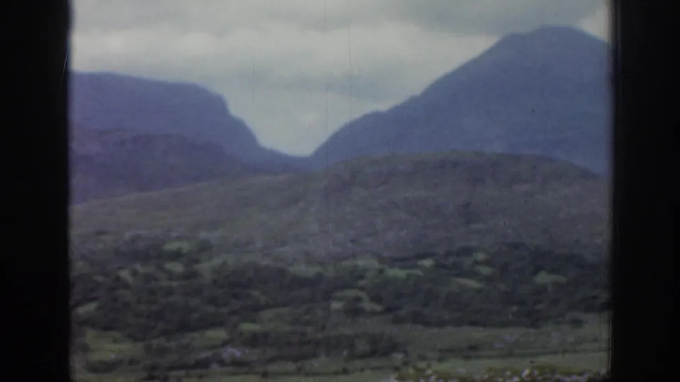 1961 Ireland landscape frame with rough green hills in the foreground and hazy mountains rising beneath an overcast sky.