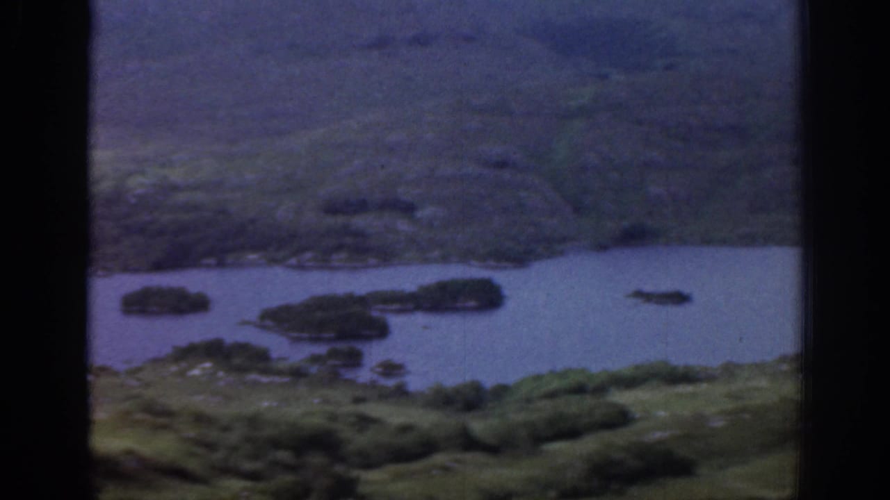 1961 Ireland 8mm frame showing a lake or broad river with small islands, green banks, and hillside terrain beyond.