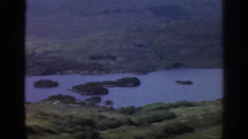 1961 Ireland 8mm frame showing a lake or broad river with small islands, green banks, and hillside terrain beyond.