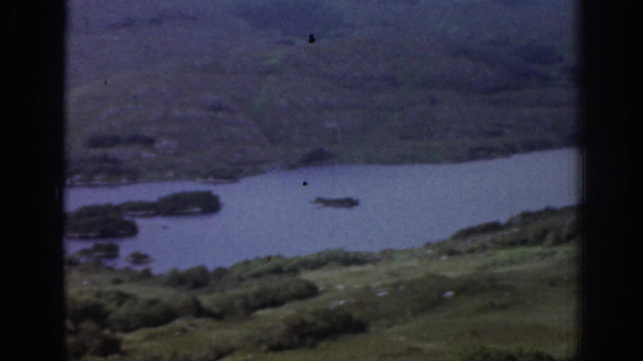 1961 Ireland 8mm view of a dark river or lake inlet between green hills, seen through heavy black frame borders.