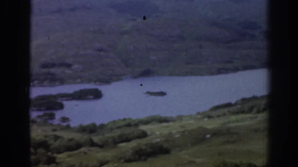 1961 Ireland 8mm view of a dark river or lake inlet between green hills, seen through heavy black frame borders.