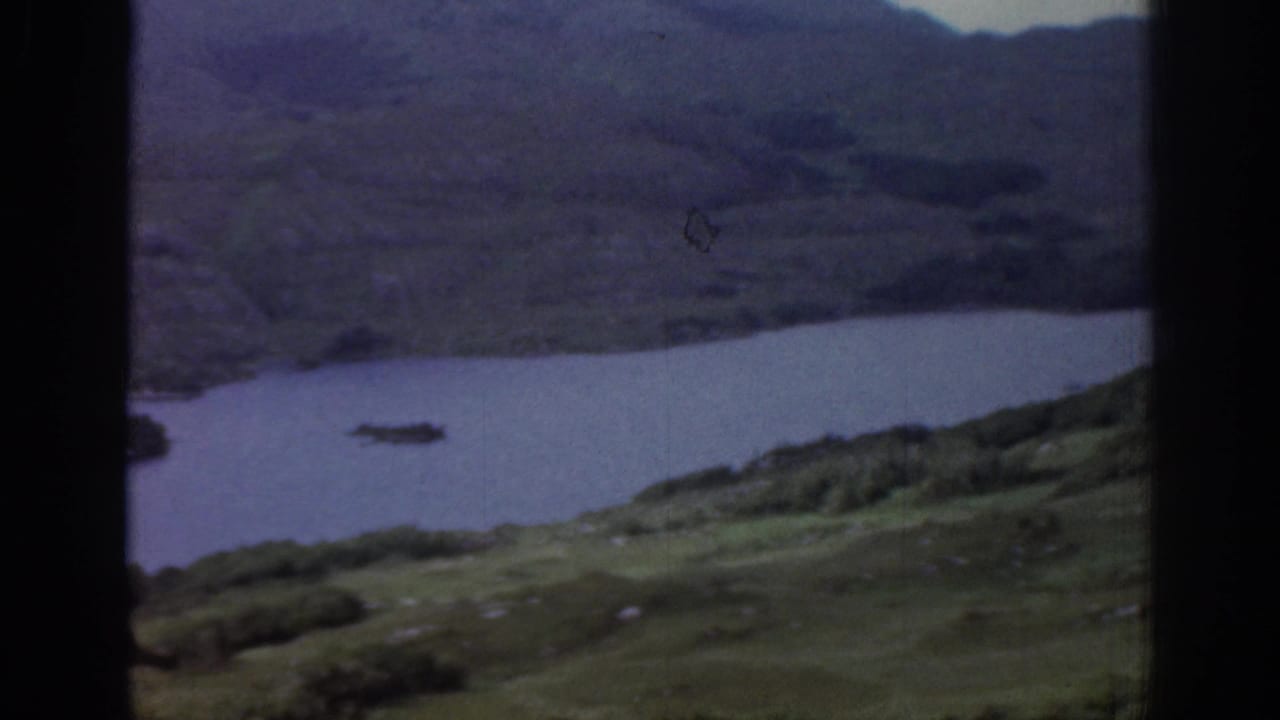 Muted 1961 Ireland home-movie frame of water below hills, with soft focus and dark vertical film borders.