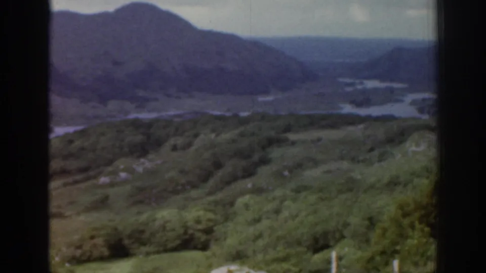Wider 1961 Ireland landscape view with green hillside vegetation, dark water, and mountains receding into the distance.