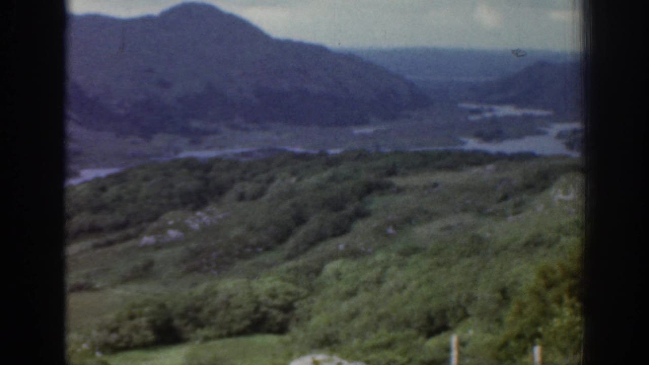 1961 Ireland home-movie frame looking across green hills toward distant mountains and winding water under cloud.