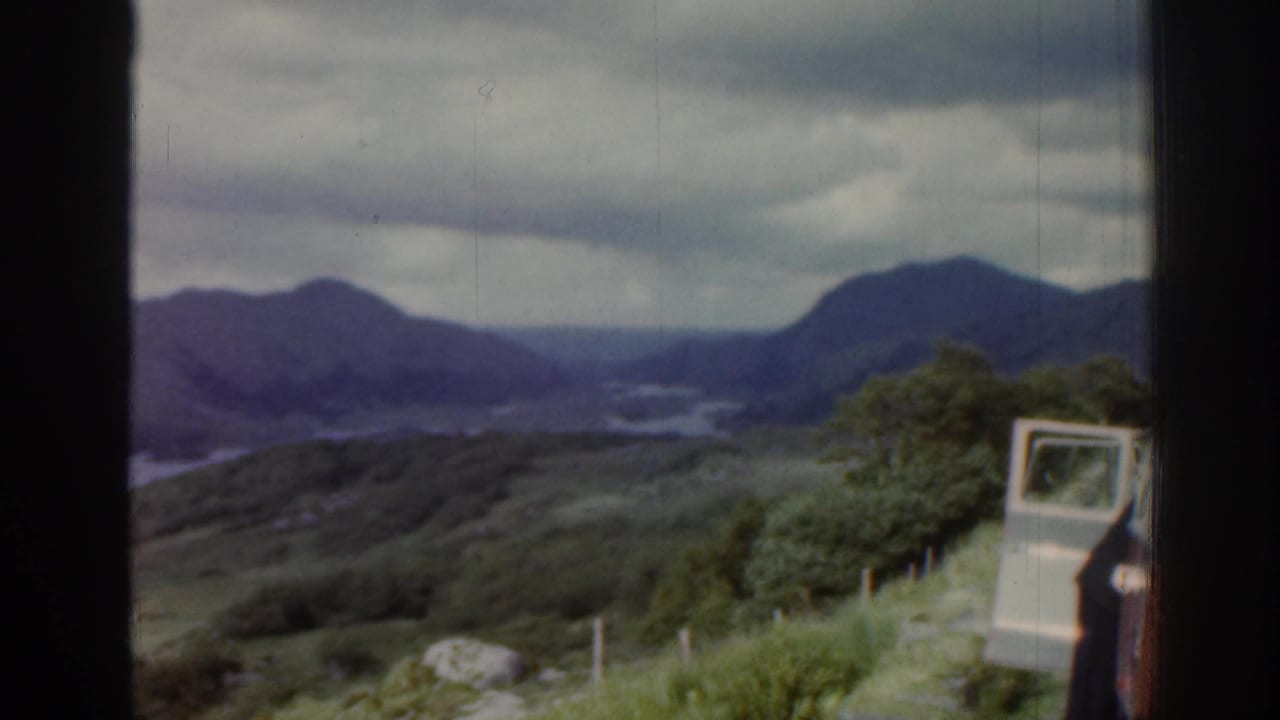 1961 Ireland roadside overlook with mountains, water, fence posts, trees, a light vehicle door and a partial person at the edge.