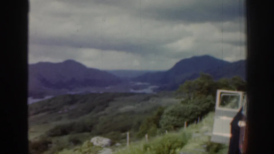 1961 Ireland roadside overlook with mountains, water, fence posts, trees, a light vehicle door and a partial person at the edge.