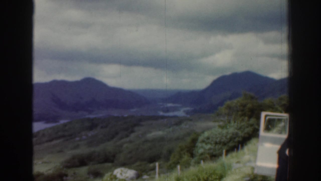 1961 Ireland 8mm frame showing a cloudy lake valley, distant mountains, grassy slope, fence posts and a small white cart-like vehicle.