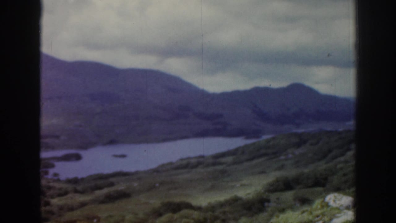 1961 Ireland 8mm frame showing a soft, muted view of lake water, green hills and distant mountains under heavy cloud cover.