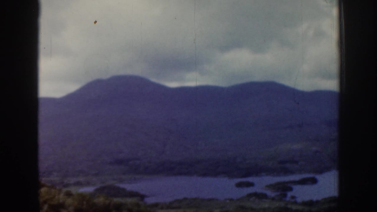 1961 Ireland 8mm frame with a closer view of rounded mountains above lake water, islands or shoreline shapes and a cloudy sky.
