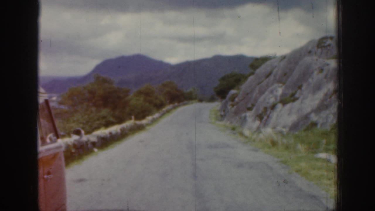 1961 Ireland road-trip frame showing a winding road, stone wall, rocky roadside, distant hills, and a partial red vehicle.