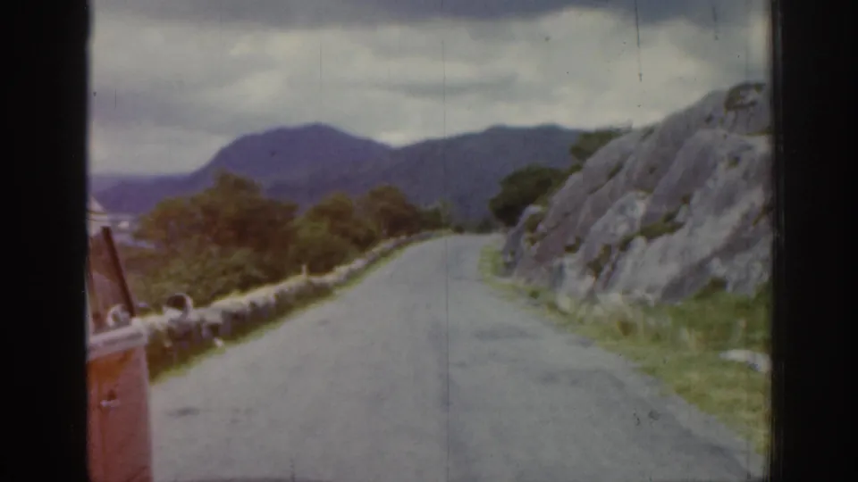 1961 Ireland road-trip frame showing a winding road, stone wall, rocky roadside, distant hills, and a partial red vehicle.